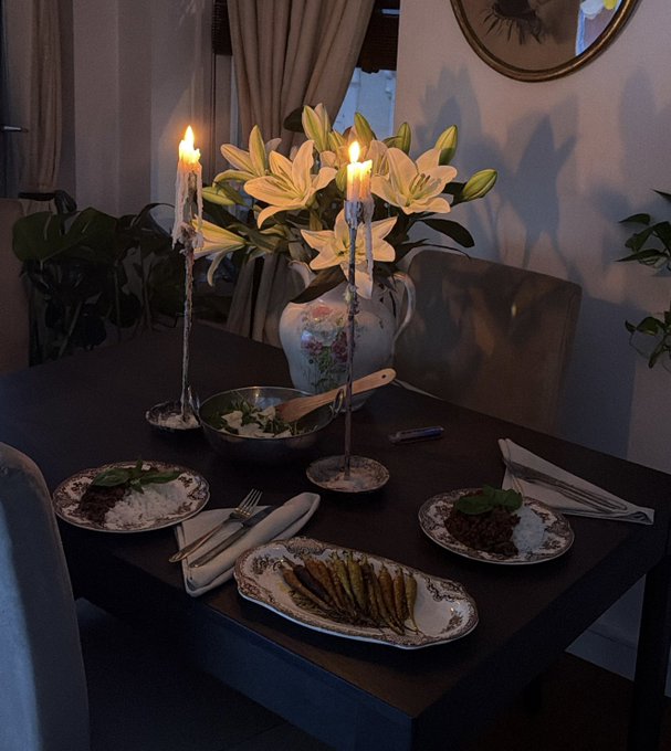 An intimate dining table setting is illuminated by lit candles, featuring a vase of lilies, two plates of food, and an asparagus dish on dark wood.