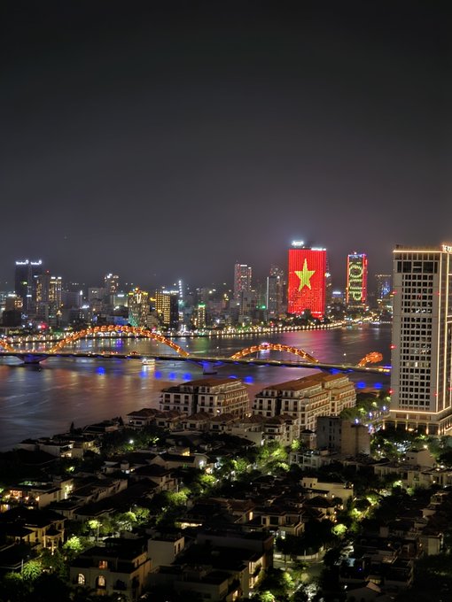 A nighttime cityscape of Ho Chi Minh City (Saigon) featuring a brightly lit bridge over the Saigon River, with skyscrapers displaying a large Vietnamese flag (red with yellow star) and "HOTEL" signage.