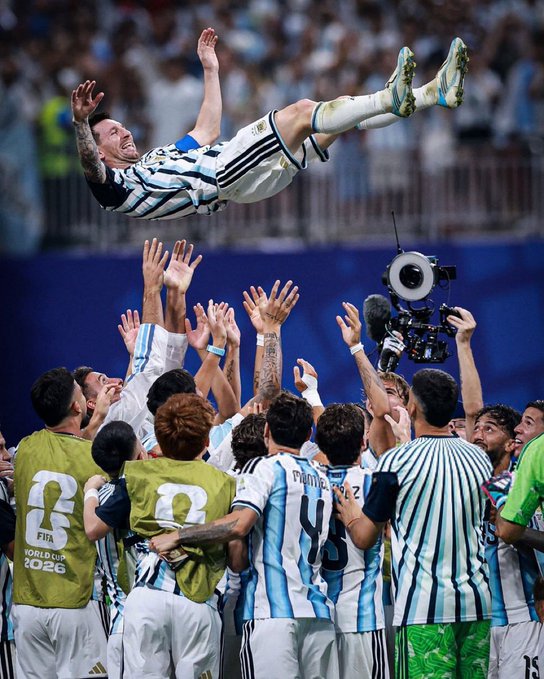 Argentine soccer player Messi being tossed in the air by teammates in light blue and white striped jerseys, celebrating a FIFA World Cup 2026 victory.