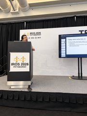 A woman presents at a podium displaying "BIOS 2026" on a stage with a screen and a stuffed animal on the floor.