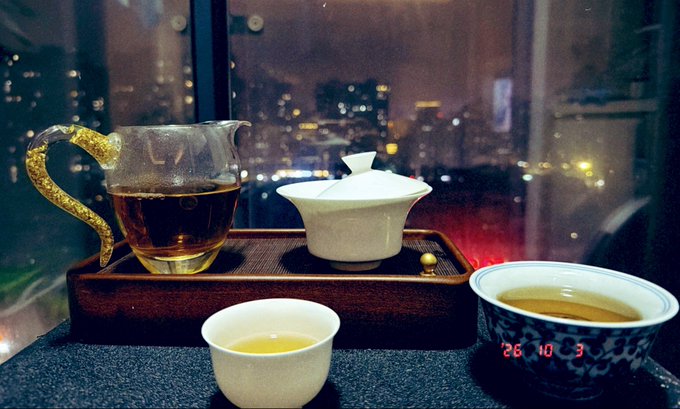 A traditional Chinese tea set on a wooden tray, featuring a glass pitcher with a gold dragon handle, a gaiwan, and two small teacups, with a cityscape visible through the window behind and a digital clock showing 26:03.