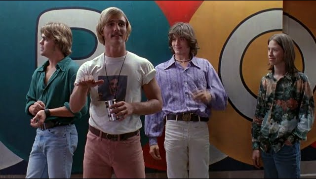 Four young men in retro 1970s clothing, including denim and floral shirts, standing together in what appears to be a film or music video still.