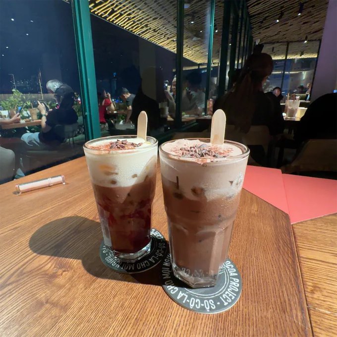 Two iced coffee drinks topped with cream and chocolate shavings, served with wooden stir sticks on coasters, sit on a wooden table in a dimly lit restaurant.