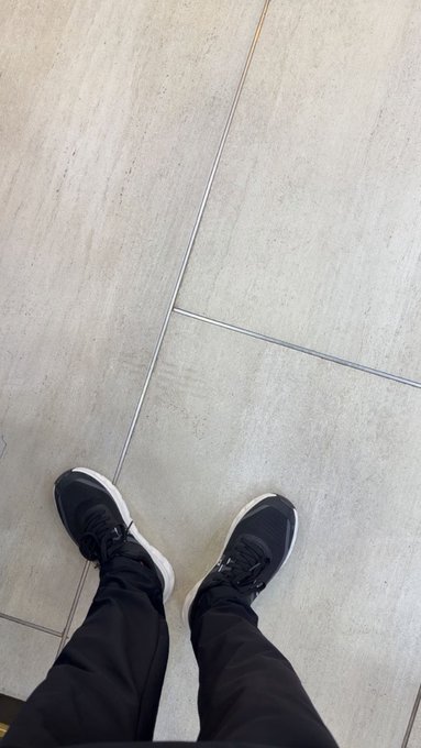 A downward view of a person's feet wearing black and white sneakers and dark pants, standing on light gray tiled flooring.