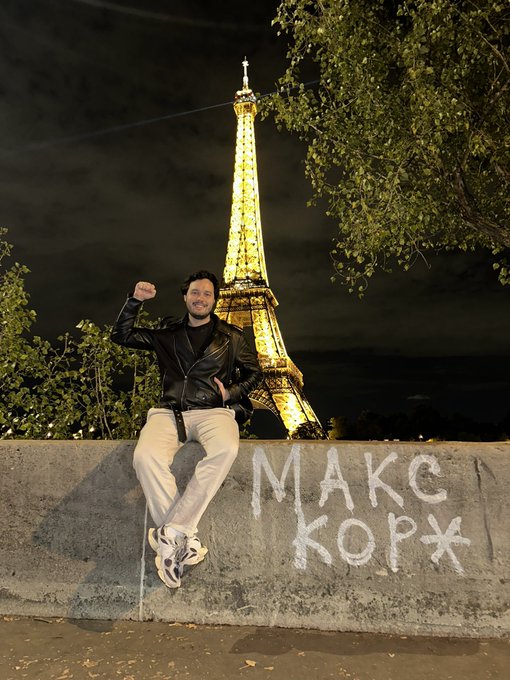A man in a black leather jacket and light pants sits on a concrete wall with white graffiti reading 'MAKE KOP*X', posing with a raised fist in front of the illuminated Eiffel Tower at night.