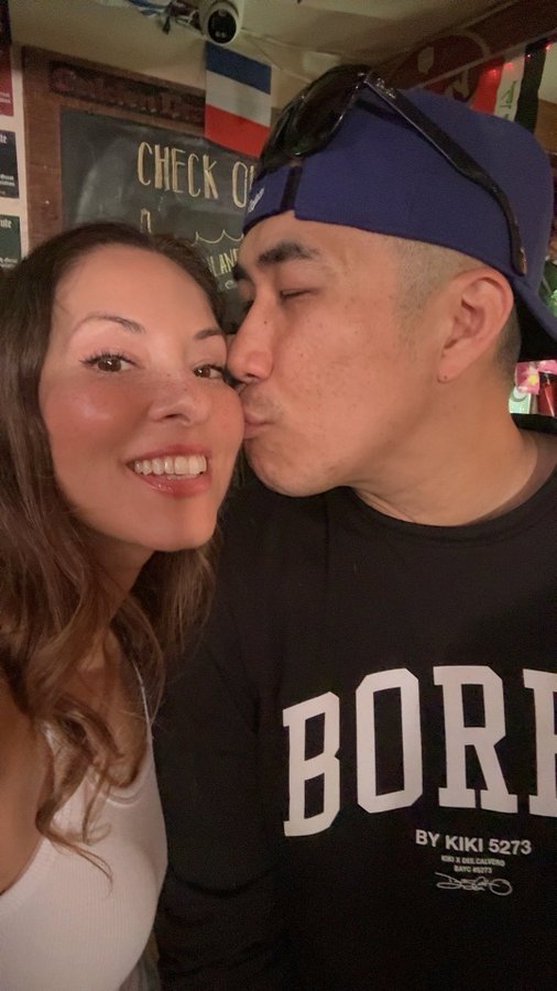 A smiling woman and a man wearing a blue backwards cap and a black 'BORN BY KIKI 5273' sweatshirt pose for a close selfie, with the man kissing her on the cheek in front of a chalkboard sign.