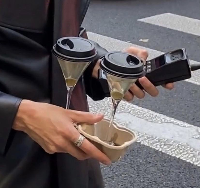 A person wearing a leather jacket holding two coffee cups with liquid pouring out into an eco-friendly container, appearing to carry a power bank or device in the same hand on a street.