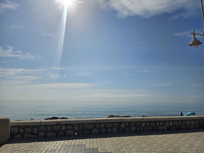 A bright, sunny ocean view from a stone-walled walkway with a street lamp visible on the right side.