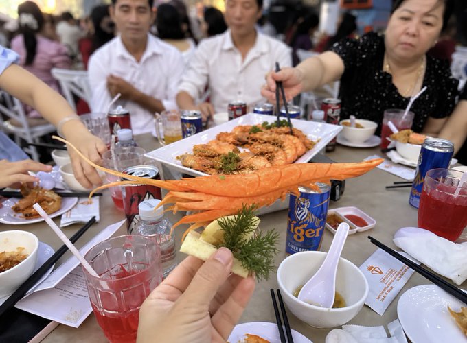 A hand holds up a massive cooked prawn at a crowded dining table filled with Tiger beer cans, plates of seafood, and various diners in what appears to be a Vietnamese restaurant.