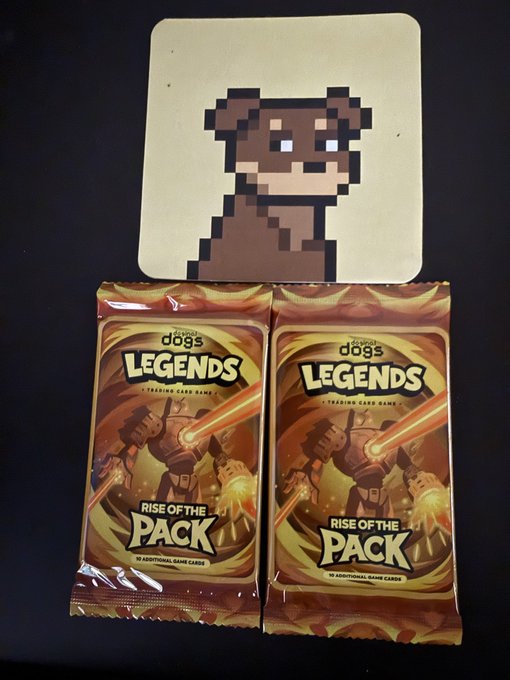 Two sealed packs of 'dogs LEGENDS - TRADING CARD GAME - RISE OF THE PACK' booster packs sit in front of a pixel-art dog coaster on a dark surface.