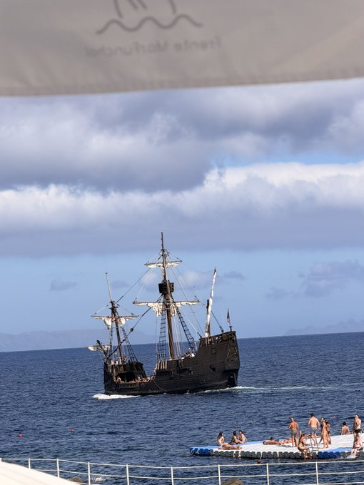 A replica galleon-style tourist ship sails on the open sea under a cloudy sky, with people enjoying an inflatable water platform nearby. The partially visible banner at the top reads "Tenerife Moun..." suggesting this is a Tenerife excursion experience. The ship's traditional tall-masted design evokes a pirate or historical voyage theme, fitting for a morning departure announcement.