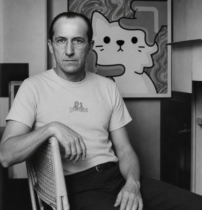 In the black and white photo, a man wearing glasses sits on a chair with his arms crossed. Behind him, there's an abstract painting of a cat that has a face and ears, giving it a cartoon-like appearance. The text "The @doodles x Elmonx official collab was so Piet Mondrian would have been very proud" is also present in the image. This suggests that the man might be a fan or admirer of the work of Piet Mondrian, who was known for his abstract art style and use of geometric shapes.
