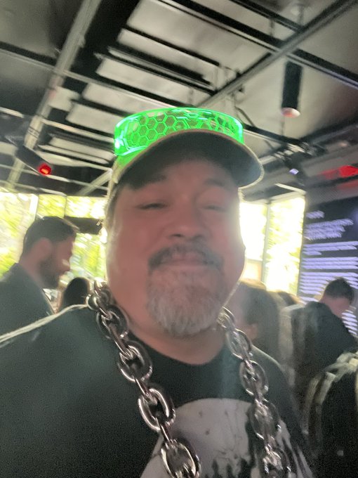 A close-up selfie of a smiling man with a goatee wearing a glowing green honeycomb visor and a large silver chain necklace in a crowded indoor space.