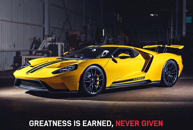 A yellow Ford GT supercar with black racing stripes and a large rear wing parked in a warehouse, with the motivational text 'GREATNESS IS EARNED, NEVER GIVEN' displayed below.