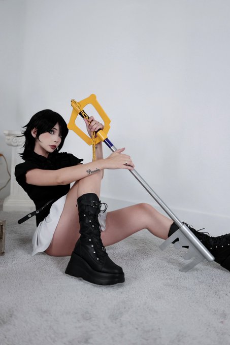 A cosplayer with short black hair, dressed in a black top and white skirt with chunky platform boots, poses on a white carpet while holding a distinctive yellow and silver Keyblade weapon. The post identifies her as "Xion," the character from Kingdom Hearts whose signature weapon is the Keyblade, fitting the cosplay's dark aesthetic and her pose with the iconic weapon.