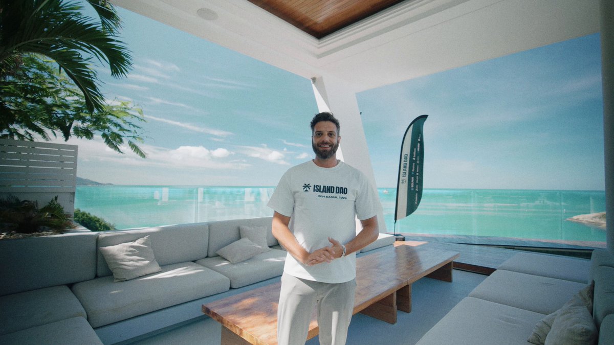 The scene shows a man standing indoors posing next to two couches. He has his arms around himself while smiling at the camera. The background features a large window overlooking an ocean view, which adds to the overall serene atmosphere of the room. There is also another person sitting on one of the couches in the image.