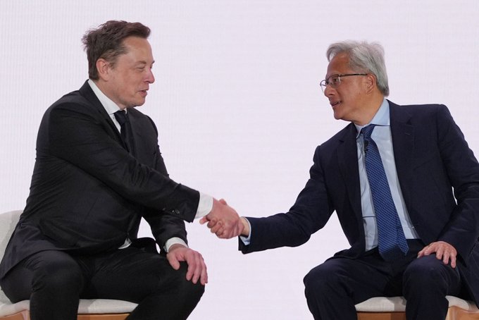 Elon Musk shakes hands with Jensen Huang, CEO of NVIDIA, while both are dressed in dark suits during a seated meeting.