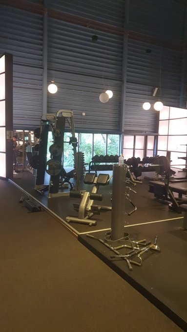 A dimly lit gym interior with various weight machines, dumbbell racks, and pendant lights hanging from a high industrial ceiling.