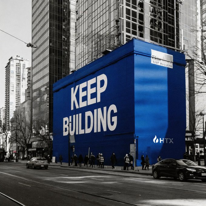 A striking urban scene features a vibrant blue building mural displaying "KEEP BUILDING" in large white letters, with the "HTX" logo visible in the corner, set against a black-and-white cityscape backdrop. The bold message echoes the post's motivational Sunday greeting, with pedestrians and traffic passing by the eye-catching installation on a city street.