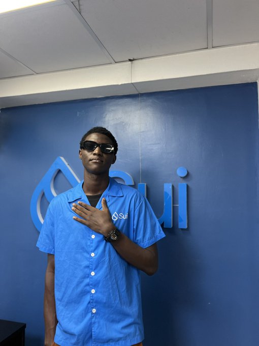 The image features a young man wearing sunglasses standing against a blue wall with a logo for "Sui Community Pluralistic Platform" written on it. The man appears to be posing for a photo, as indicated by his hand raised above his head in front of him.