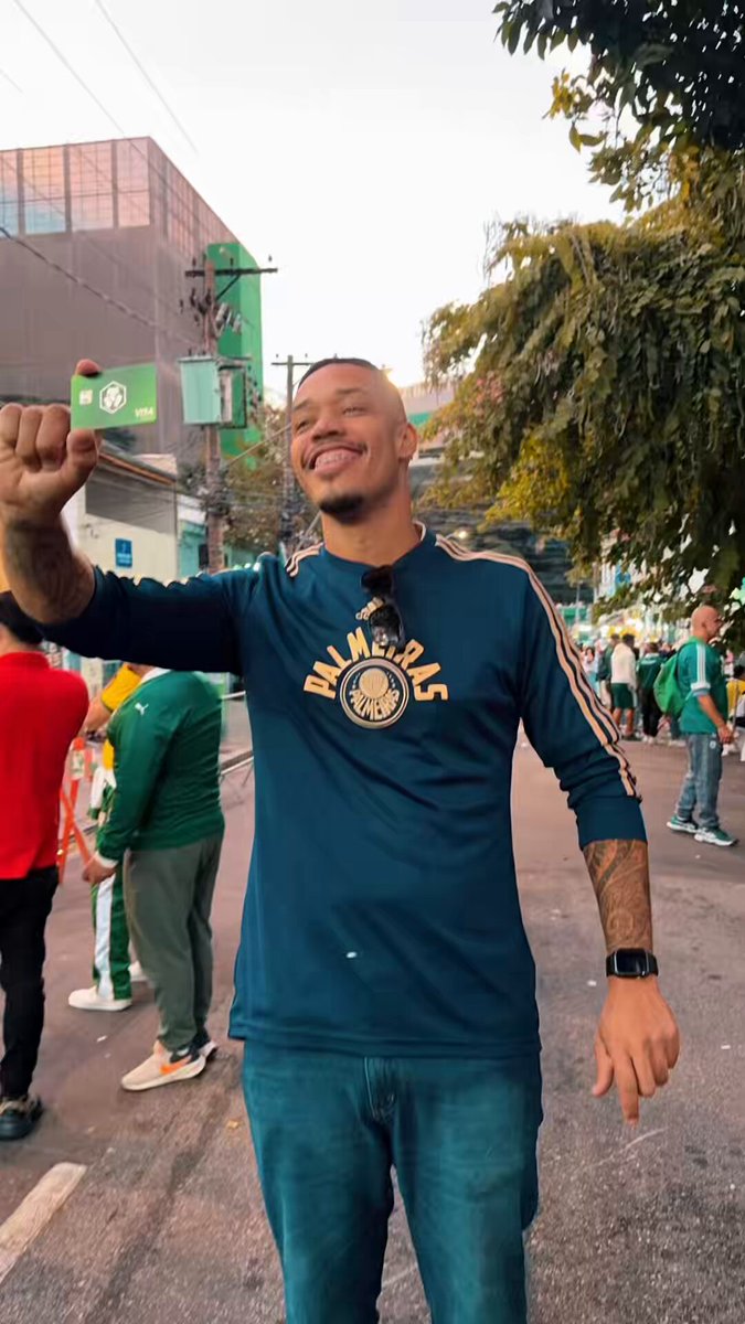 The image features a man wearing a blue shirt with gold lettering standing on a street corner holding up his hand. The background shows other people walking around, indicating that they are likely at an event or gathering. There is also a green sign visible in the scene.