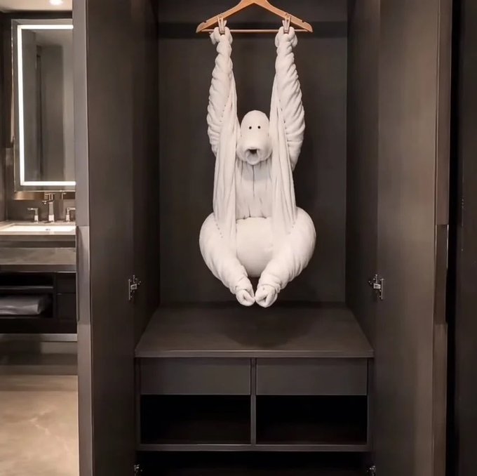 A white towel art creation shaped like a dog/animal hanging from a wooden hanger inside a dark gray closet, next to a bathroom vanity with mirror.