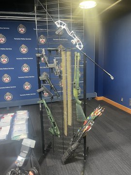 A display at Toronto Police Service headquarters shows compound bows and arrows arranged on a stand, with "Toronto Police Service" branding visible on the backdrop. The weapons exhibit relates to the investigation where a man was allegedly abducted at gunpoint near Finch Avenue and Bayview Avenue and forced to surrender access to his cryptocurrency.