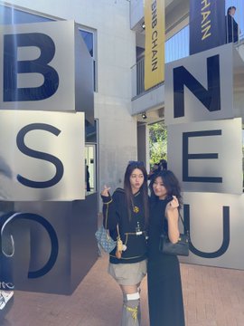 Two young women pose with peace signs in front of a building displaying large letters spelling 'BSEND' and 'REALM', with 'SEIKAN' banners visible above.