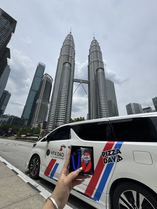The image shows a person holding up a small object that appears to be a wallet or cellphone case with a picture of a pizza on it. The background features two tall buildings, possibly the Petronas Twin Towers, which are iconic landmarks in Kuala Lumpur, Malaysia. The person is standing next to a white car parked nearby.