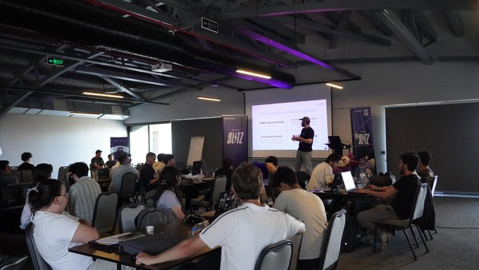 A presenter at the Monad Blitz Ankara v3 hackathon stands before an audience of attendees seated at tables with laptops, with the event branding "Blitz" visible on banners flanking the projection screen. The single-day IRL hackathon brought builders together to develop on Monad, with the event showcasing projects like WICK, an AI-powered market maker hook inspired by Uniswap v4.