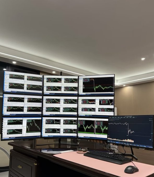 A professional trading workstation featuring multiple monitors arranged on a wall, displaying various financial charts and graphs, with a keyboard and mouse setup on a desk below.