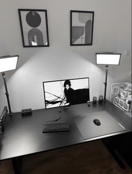 A minimalist desk setup includes a computer monitor displaying an anime character, two LED light panels, framed wall art, a mechanical keyboard, and a mouse on a dark gray desk.