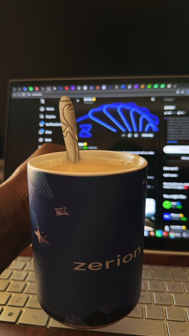 A hand holding a dark blue 'zerion' branded mug with a spoon in it, set against a laptop keyboard and screen displaying a crypto portfolio with a blue dinosaur logo.
