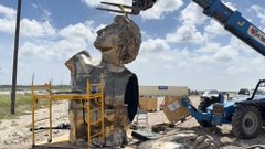 Construction workers on scaffolding assemble a large stone bust statue at an outdoor worksite with a crane in the background.