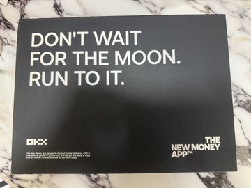 A black advertisement poster on a marble surface with the text 'DON'T WAIT FOR THE MOON. RUN TO IT.' along with the OKX logo and the tagline 'THE NEW MONEY APP'.