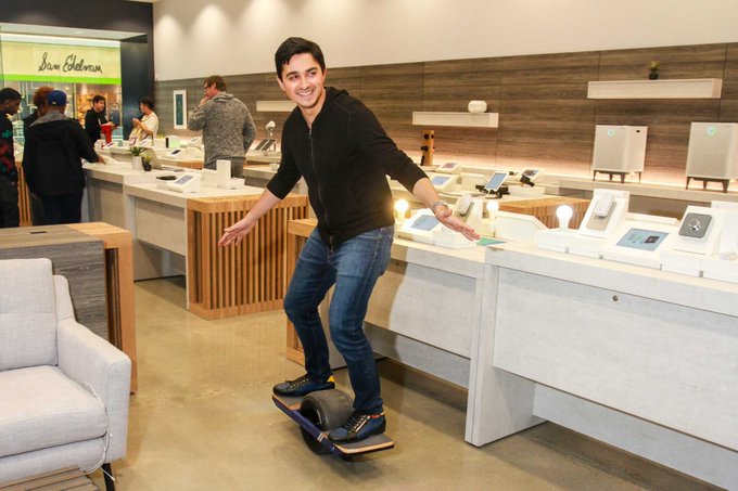A smiling man balances on a small one-wheeled electric skateboard inside a brightly lit electronics store, with various tech devices displayed on counters behind him.