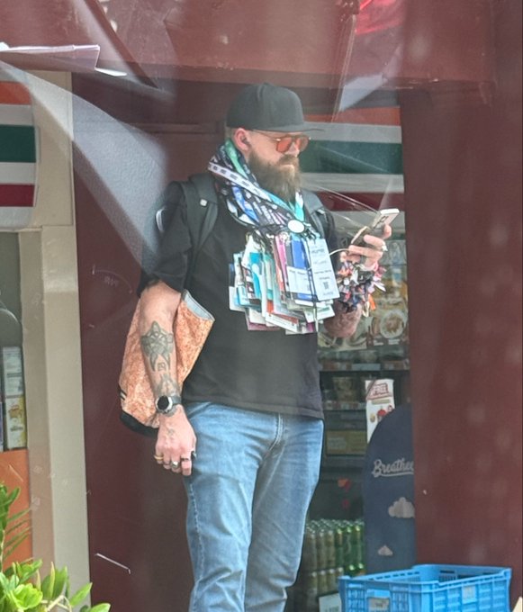 A bearded man wearing a black cap, orange-tinted sunglasses, and a lanyard covered in colorful event badges looks down at his phone while holding a water bottle, with a "7-Eleven" sign and "Breathes" branded cooler visible in the background. The image captures @Player1Taco, a well-connected crypto community member spotted at conferences like Chainlink NYC, illustrating the post's advice that real crypto "alpha" comes from meeting passionate, badge-laden attendees in person at events.