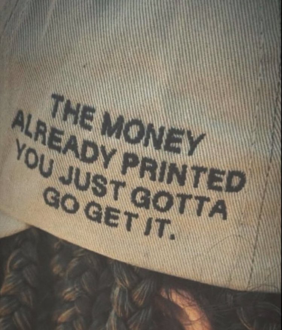 A close-up photo of a canvas item (possibly a bag or hat) printed with the text 'THE MONEY ALREADY PRINTED YOU JUST GOTTA GO GET IT.'