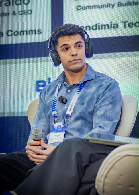 A man wearing headphones and a lanyard labeled "Scalyn" sits at what appears to be a conference panel, with partial signage for "Blockchain Rio" and "Community Builder" visible behind him. The image aligns with the post's caption, which thanks Blockchain Rio for the opportunity to participate in the event, highlighting the networking and learning experience at the conference.