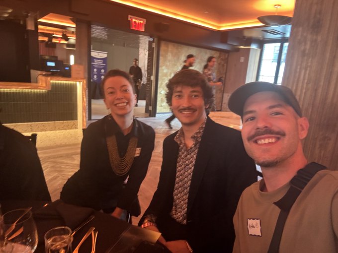 A selfie of three people smiling in what appears to be a restaurant or hotel lobby, including a woman in a black uniform with gold chains, a man in a patterned shirt and blazer, and a man in a cap and tan shirt in the foreground.