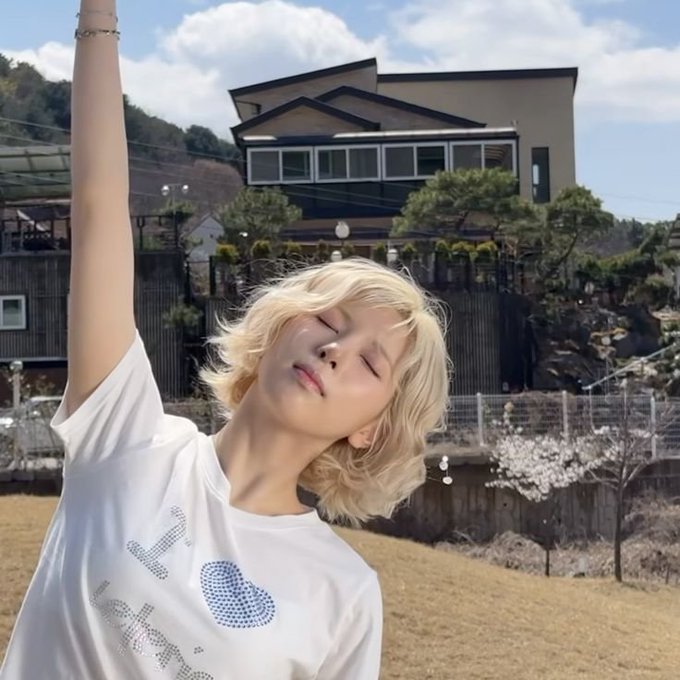A young person with blonde, wavy hair posing outdoors with one arm raised, wearing a white t-shirt with blue graphics, set against a background of a house and blossoming trees.