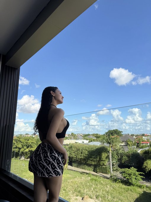 A woman with long curly hair wearing a black top and patterned shorts stands at a window looking out at a landscape with houses, trees, and a partly cloudy blue sky.