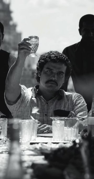 Black and white photo of a man with a mustache raising a glass while seated at a table with other people in the background.