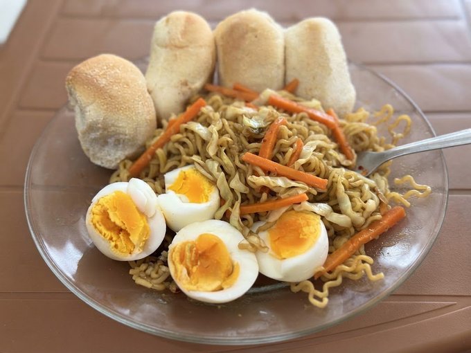 A plate of stir-fried noodles with shredded carrots and cabbage, served with hard-boiled eggs cut in half and several bread rolls.