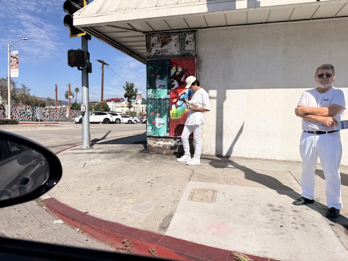 A street scene shows two people dressed in all white near a building corner, with one person looking at a phone, photographed from inside a vehicle.