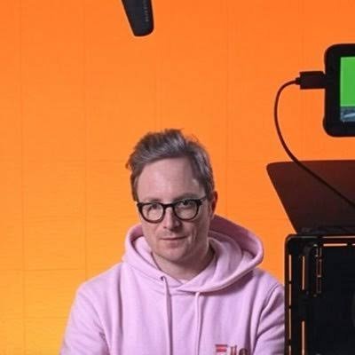 Portrait of a man with glasses wearing a pink hoodie, seated in front of an orange backdrop with video production equipment visible.