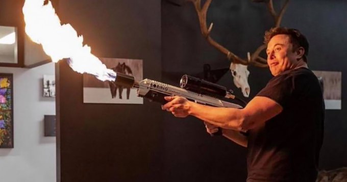 Elon Musk is shown holding a large futuristic rifle that is firing a burst of flame, in a room decorated with antlers and wildlife photos.