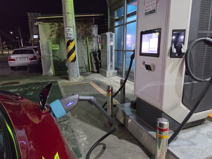 Nighttime photograph of an electric vehicle (blue Tesla) plugged into a Korean DC fast charging station, with Korean text visible on the station signage.