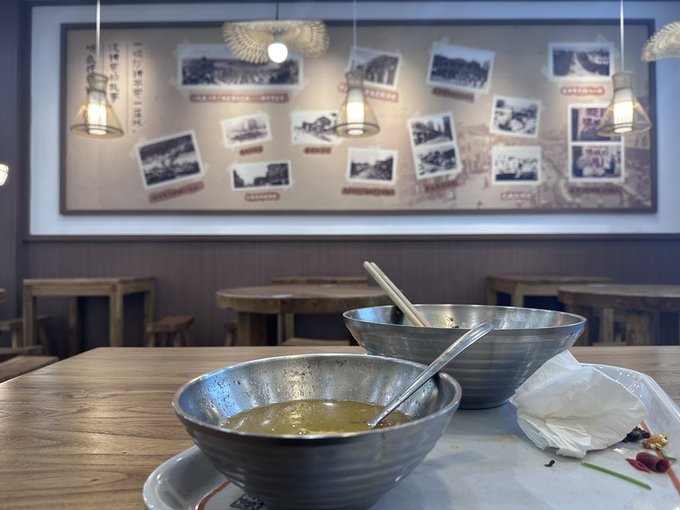 Two nearly empty metal bowls sit on a wooden table in a restaurant, with a decorative photo wall and pendant lights visible in the background.