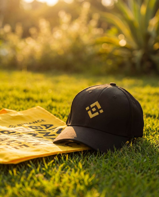 A black Binance-branded baseball cap rests on a yellow Binance merchandise bag atop a sunlit grassy lawn, tying into the "GM早！" (Good morning!) post by evoking a relaxed, ready-to-go vibe perfect for discussing the week's crypto plans.